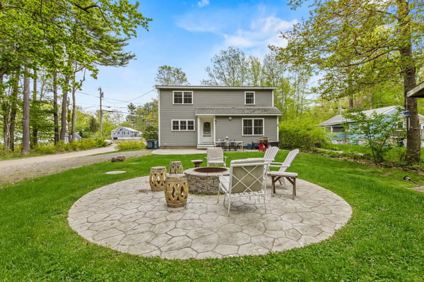Two-story gray house with patio fire pit, Adirondack chairs, and stone stools surrounded by tall trees