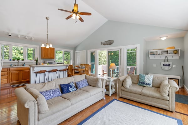 Open-plan living room with two beige sofas, hardwood floors, vaulted ceiling with ceiling fan, and kitchen with wood cabinets visible in background