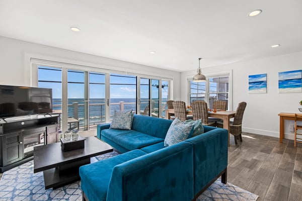 Open-plan living room with blue sectional sofa, ocean view through floor-to-ceiling windows, and dining area with wicker chairs.
