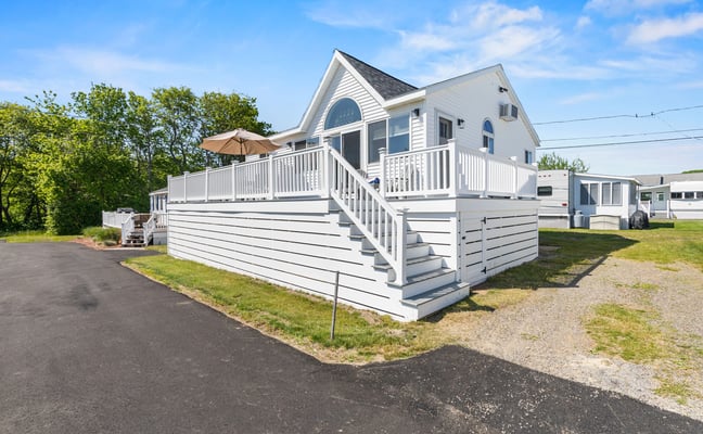 White elevated vacation home with wraparound deck, umbrella, and stairs, surrounded by green lawn and trees